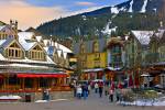 Stock photo of pedestrian activity and shops along the Village Stroll in Whistler Village, British Columbia, Canada. The tall rooftops are mostly snow covered as are the mountains in the background.