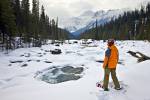 Stock photo of a woman wearing snow shoes looking at the snow covered winter landscape surrounding the Mistaya River with Mount Sarbach (3155 metres/10351 feet) in the background in Mistaya Canyon along the Icefields Parkway in Banff National Park, Canadi