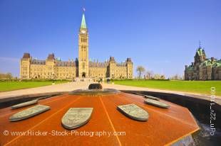 Stock photo of the Centre Block Parliament Buildings and Peace Tower against a clear blue sky in the background. A close up of the Centennial Flame monument makes up the entire foreground in this historic scene.