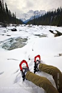 Stock photo of a person wearing snowshoes sitting on the snow covered banks of the Mistaya River during winter with Mount Sarbach in the background, Mistaya Canyon, Icefields Parkway, Banff National Park, Canadian Rocky Mountains, Alberta, Canada.