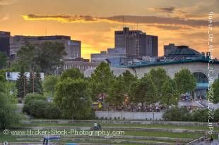 Stock photo of The Forks National Historic Site at sunset in the City of Winnipeg, Manitoba, Canada. You can see the Market and Marina as well as tall city buildings against an orange-yellow sky with clouds lined by sunlight.